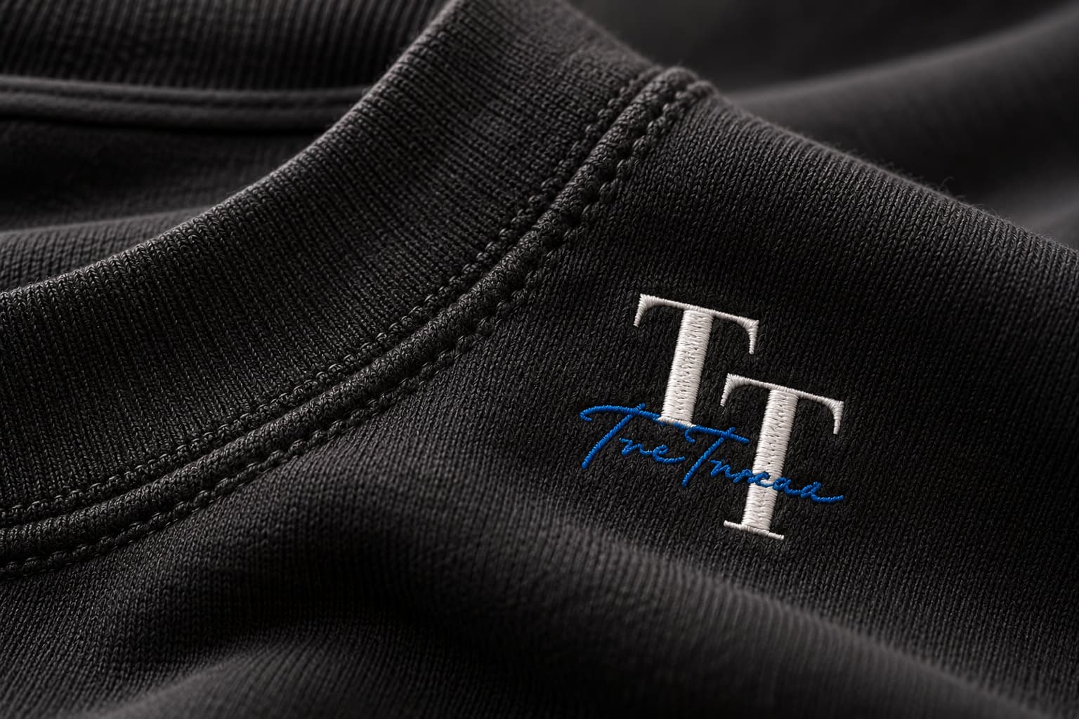 Close-up of an embroidered TreTrove monogram beside a stitched neckline on black fabric